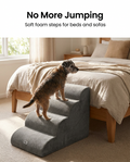 Small dog climbing the four step grey foam pet stairs beside a bed